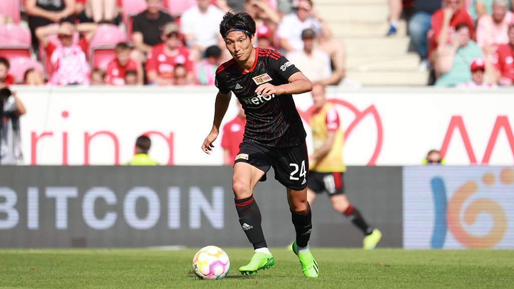 Ein Fußballspieler in einem schwarzen Trikot mit roten Akzenten dribbelt den Ball auf einem grünen Spielfeld. Fans sind im Hintergrund zu sehen.