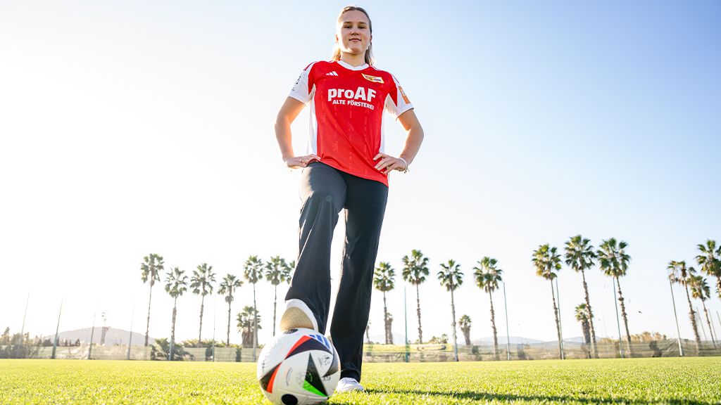 An athlete in a red jersey stands on a soccer field with a soccer ball in front of her. Palm trees are visible in the background.