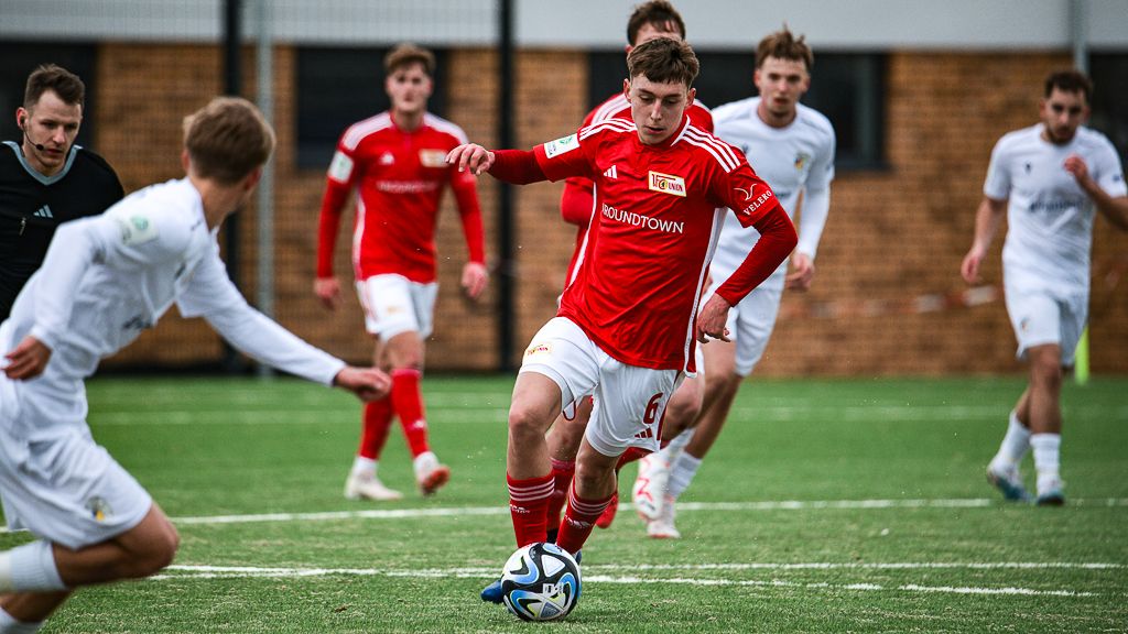 Ein Spieler in rotem Trikot dribbelt einen Ball auf einem Fußballfeld, während andere Spieler im Hintergrund aktiv sind.