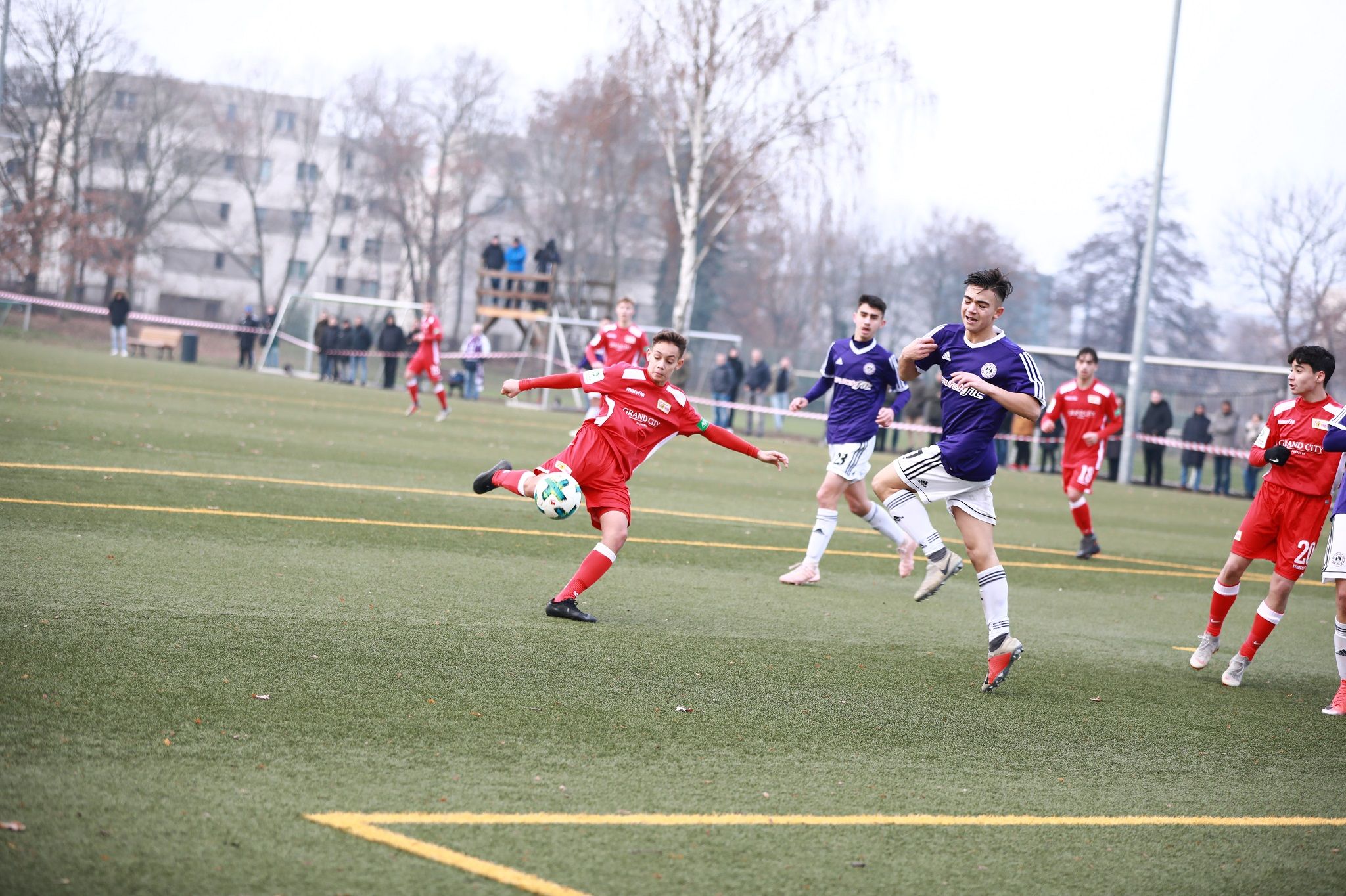 Ein Jugendfußballspiel auf einem Rasenplatz, mit Spielern in roten und lila Trikots, während ein Spieler einen Schuss ausführt.