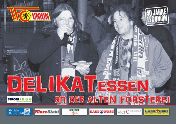 Zwei Männer in Fußballschals stehen lächelnd in einer urbanen Umgebung, umgeben von Werbung für eine Veranstaltung.