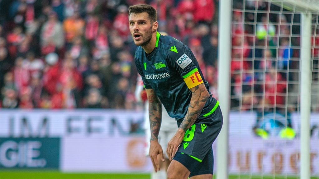 Ein Fußballspieler mit Kapitänsbinde in grünem Trikot steht auf dem Platz und schaut angespannt in die Ferne.