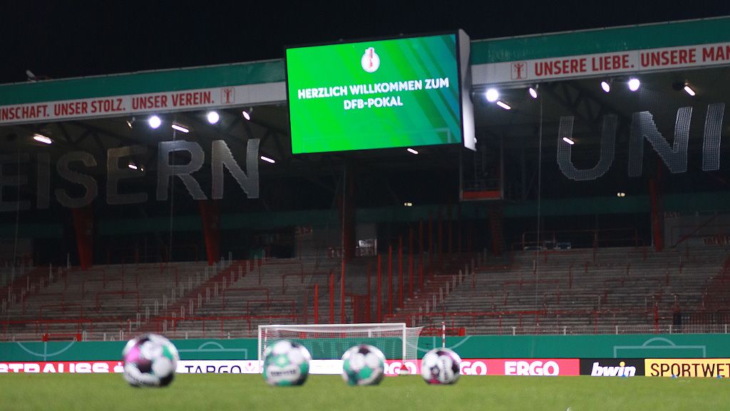 Beleuchtetes Stadion mit einem Bildschirm, der "Herzlich Willkommen zum DFB-Pokal" anzeigt, und mehreren Fußbällen im Vordergrund.