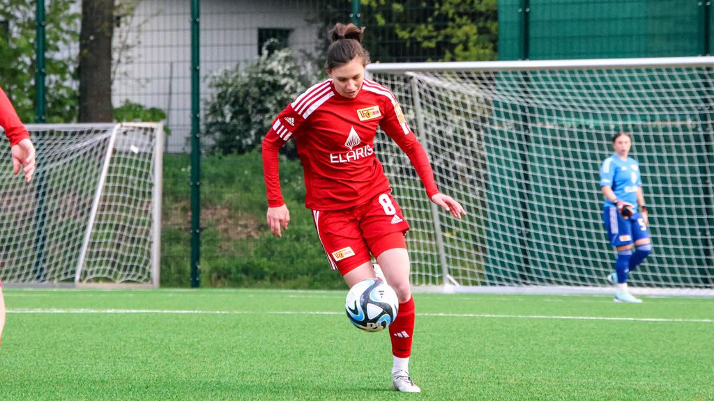Spielerin in rotem Trikot mit der Nummer 8 dribbelt den Ball auf einem Fußballplatz. Im Hintergrund ein Tor und eine andere Spielerin.