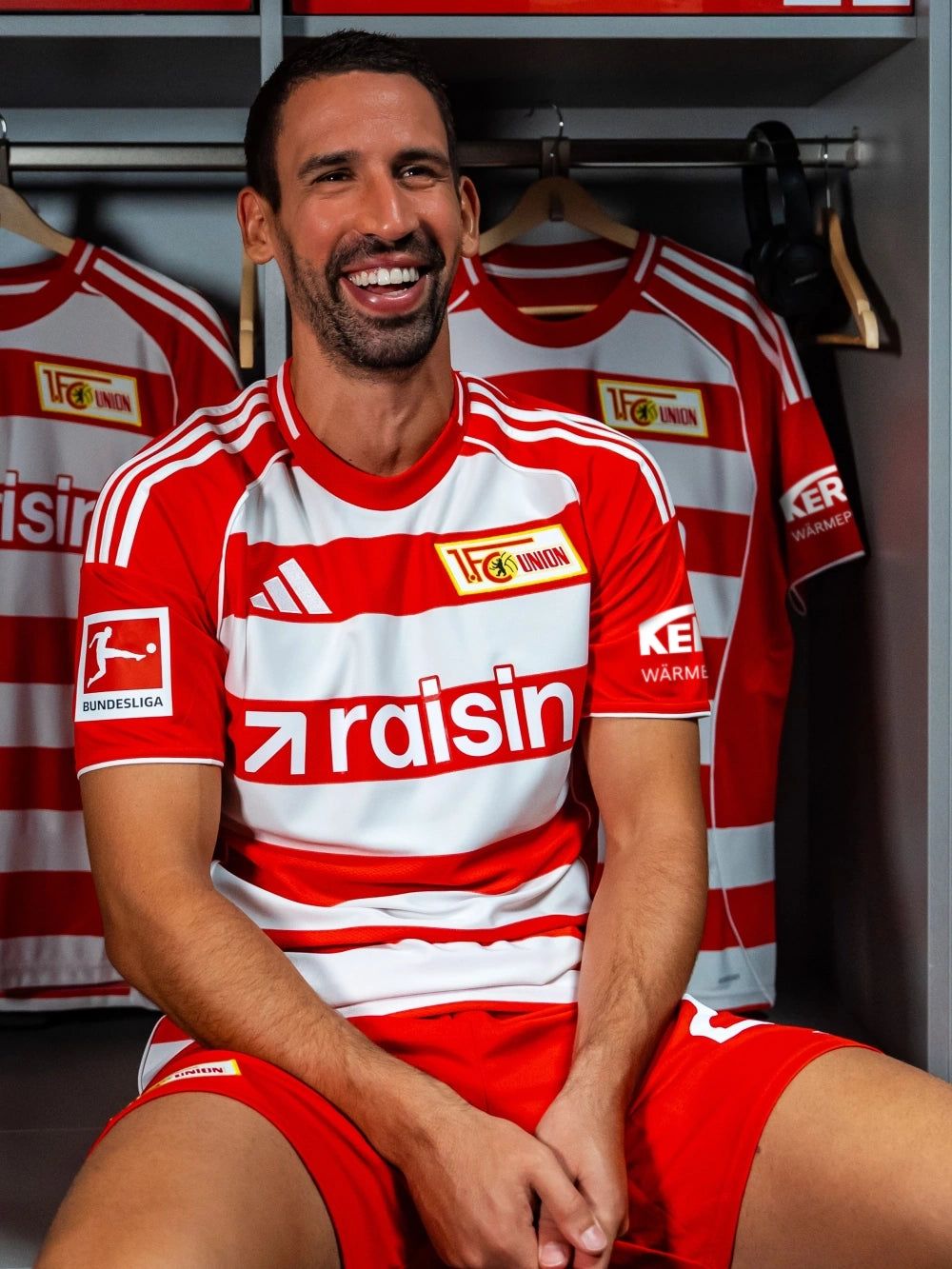 Lächelnder Fußballspieler in rot-weißem Trikot sitzt in der Umkleidekabine, umgeben von weiteren Trikots.