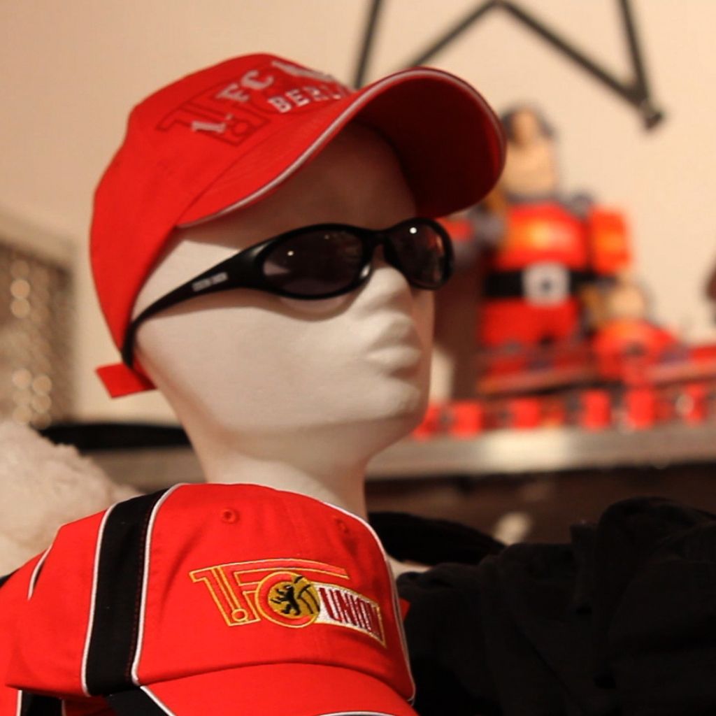 Schaufensterpuppe mit rotem Baseballcap und Sonnenbrille, hinter ihr Spielzeuge in bunten Farben.