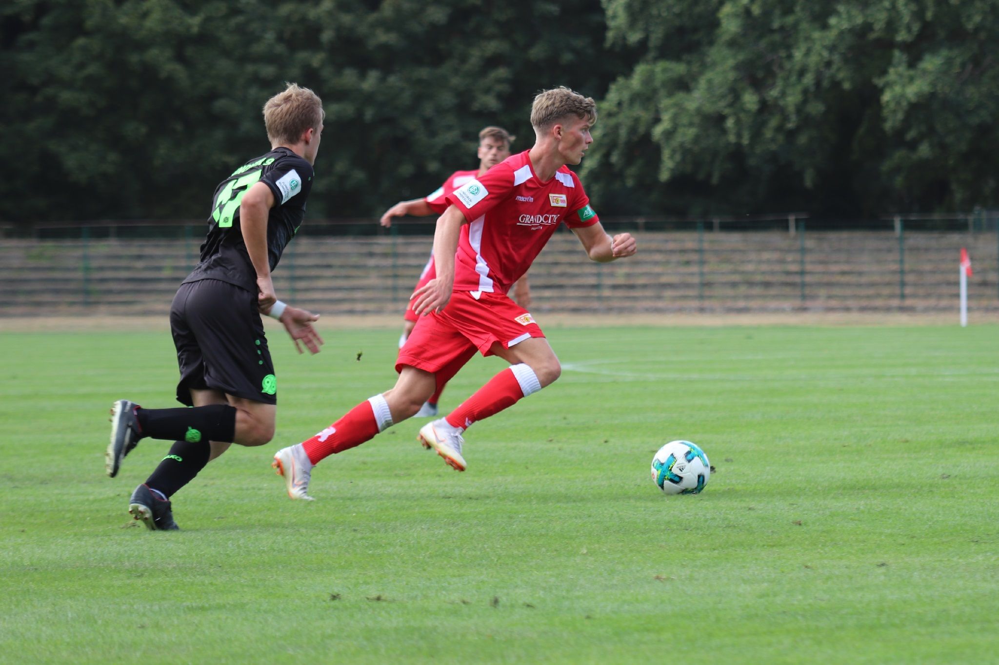 Ein Fußballspieler in rotem Trikot rennt mit dem Ball, während ein gegnerischer Spieler in Schwarz ihn verfolgt.