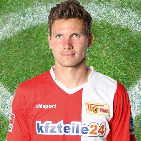 Ein Fußballspieler trägt ein rotes Trikot mit dem Logo des Vereins und steht vor einem grünen Fußballfeld.