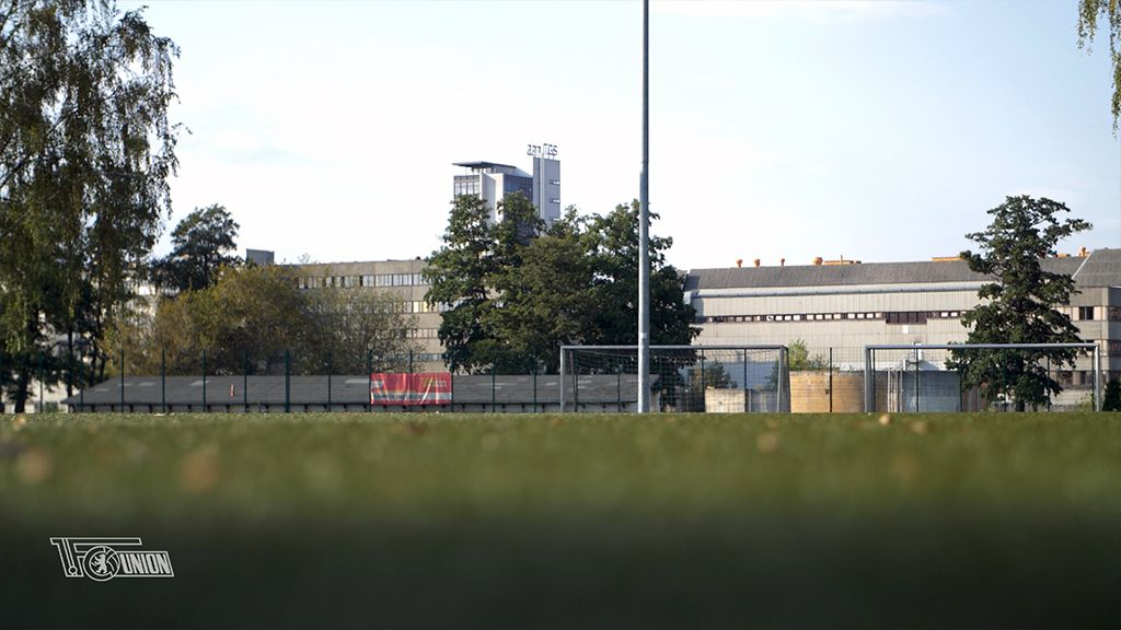 Grünfläche mit einem Fußballtor im Vordergrund und einer modernen Gebäudefassade im Hintergrund unter blauem Himmel.