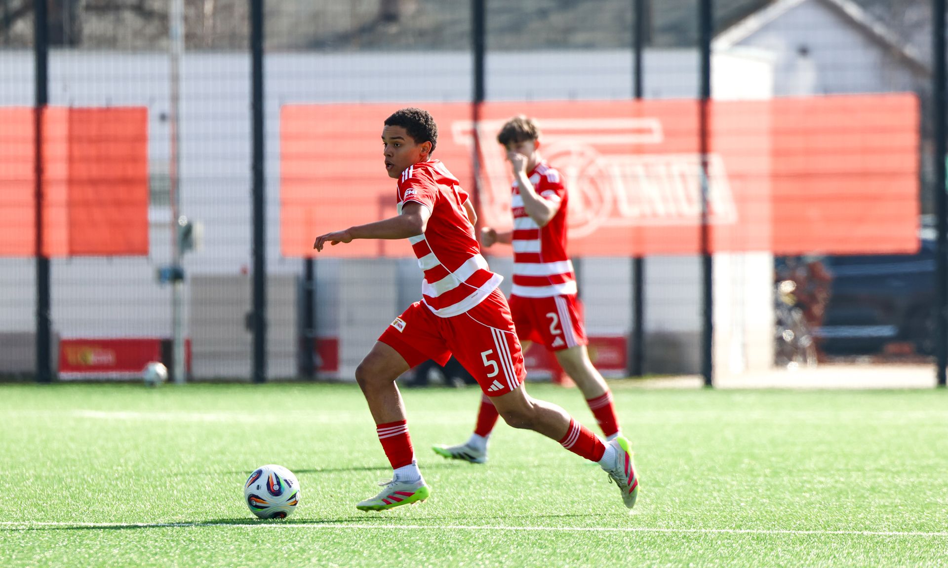 Spieler in roter Ausrüstung läuft mit dem Ball über ein Fußballfeld, während ein Teamkollege im Hintergrund steht.