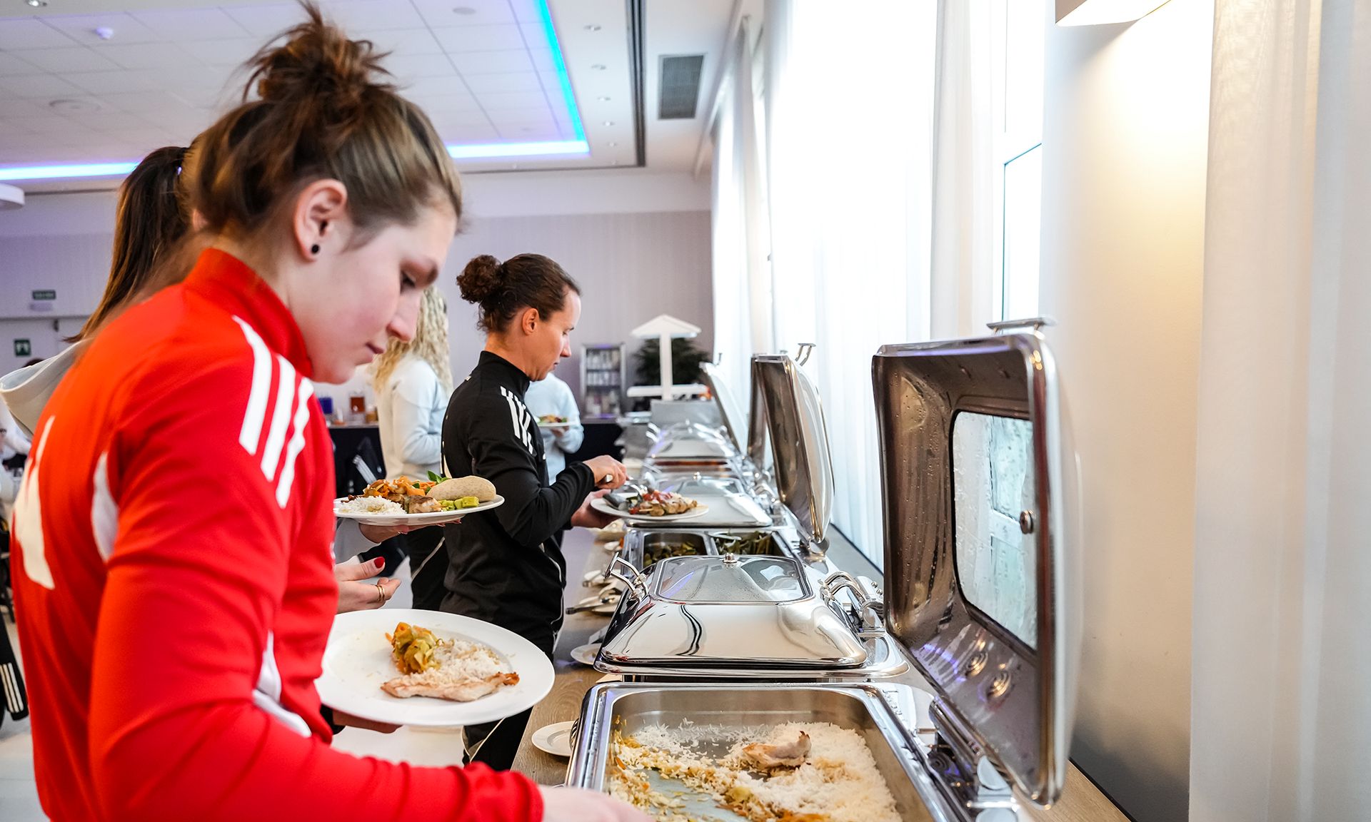Personen in Sportkleidung bedienen sich an einem Buffet mit verschiedenen Speisen. 