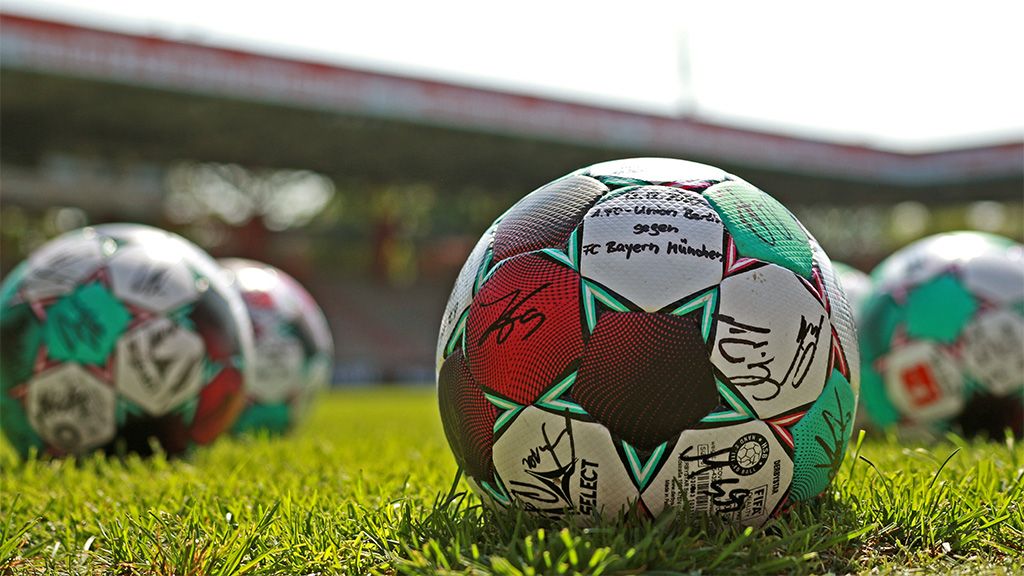 Ein signierter Fu ßball liegt auf einem grünen Rasenfeld, im Hintergrund sind verschwommene Stadionstrukturen zu sehen.