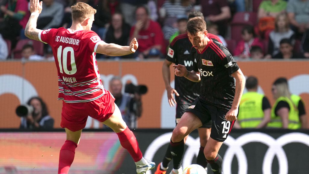 Two soccer players competing for the ball during a game in a stadium. Spectators in the stands in the background.