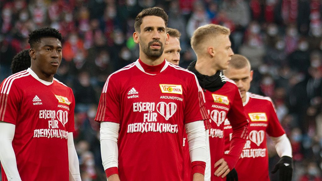 Spieler einer Fußballmannschaft in roten Trikots mit der Aufschrift „EISERN FÜR MENSCHLICHKEIT“ stehen auf dem Feld.