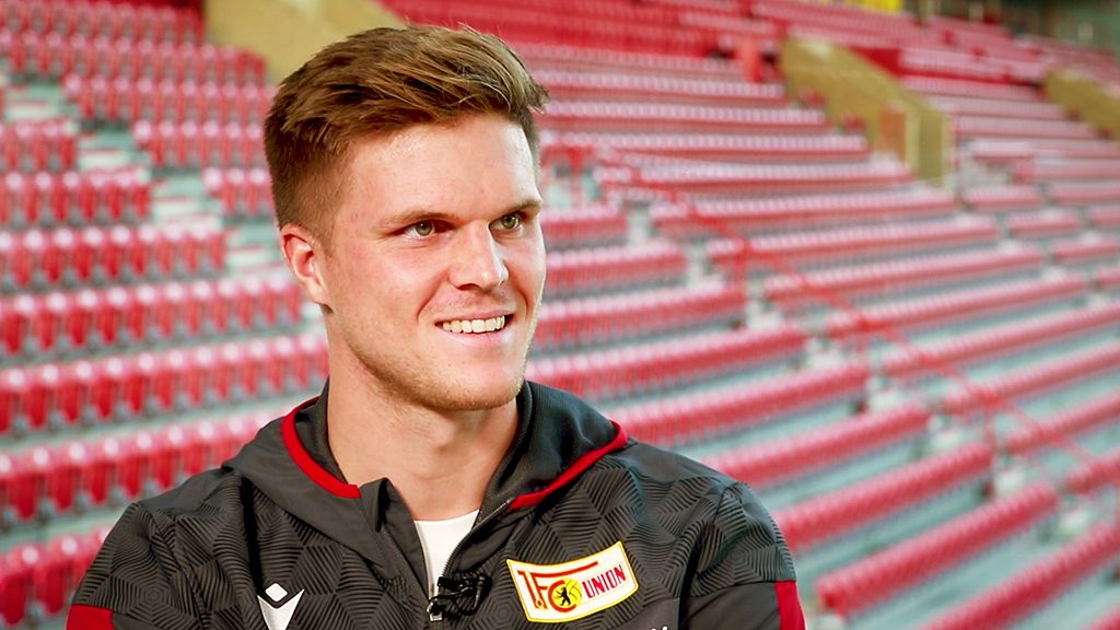 A young man with brown hair smiles in an empty stadium, dressed in a tracksuit with a club logo.
