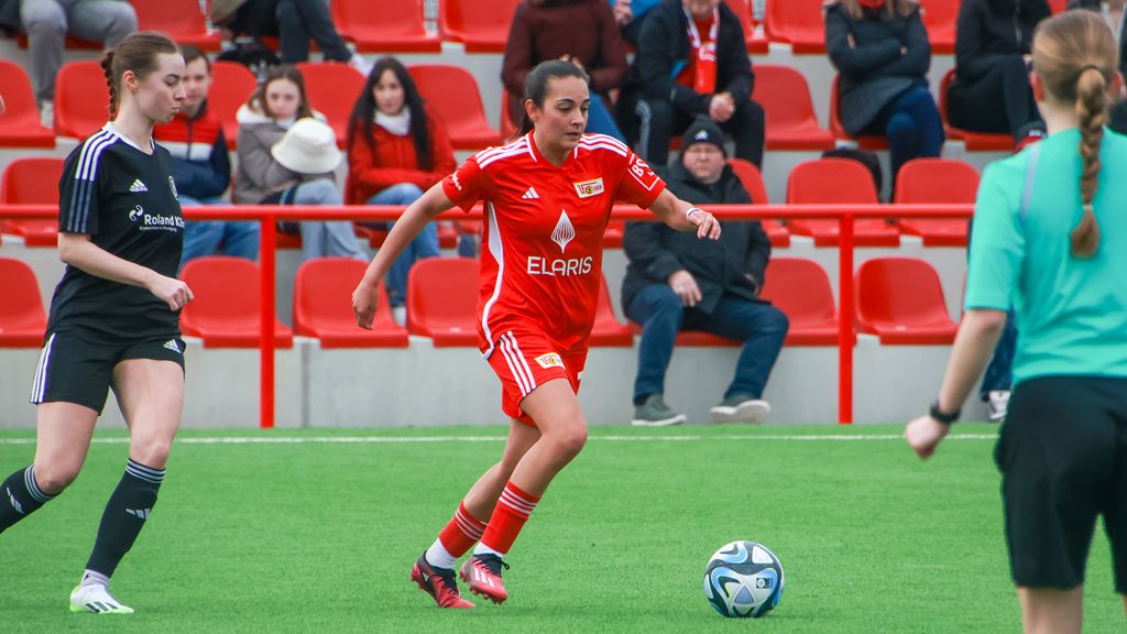 Game scene in women's football: A player in a red jersey dribbles the ball on a field, while other players stand in the background.