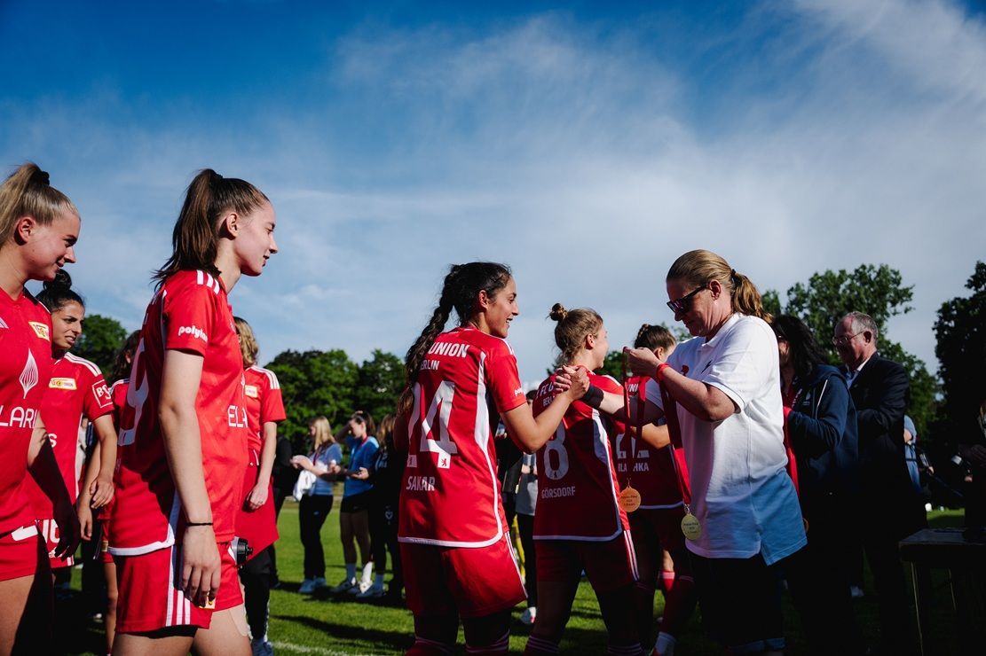 Mädchenmannschaft in roten Trikots erhält Medaillen bei einer Siegerehrung auf einem Sportplatz.