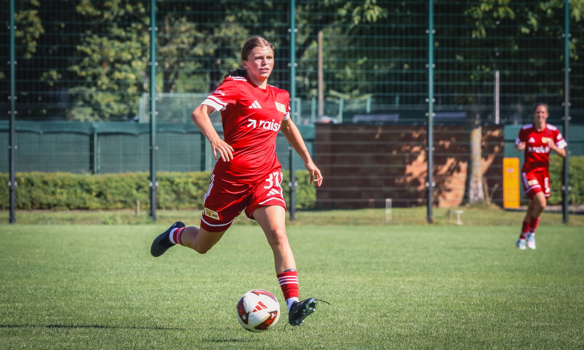 Eine Fußballspielerin in rotem Trikot läuft mit dem Ball über ein grünes Spielfeld. Im Hintergrund ist ein weiteres Teammitglied zu sehen.