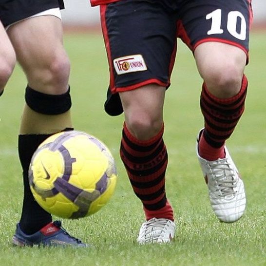 Zwei Fußballspieler stehen nebeneinander, während ein gelber und lila Fußball zwischen ihnen rollt.