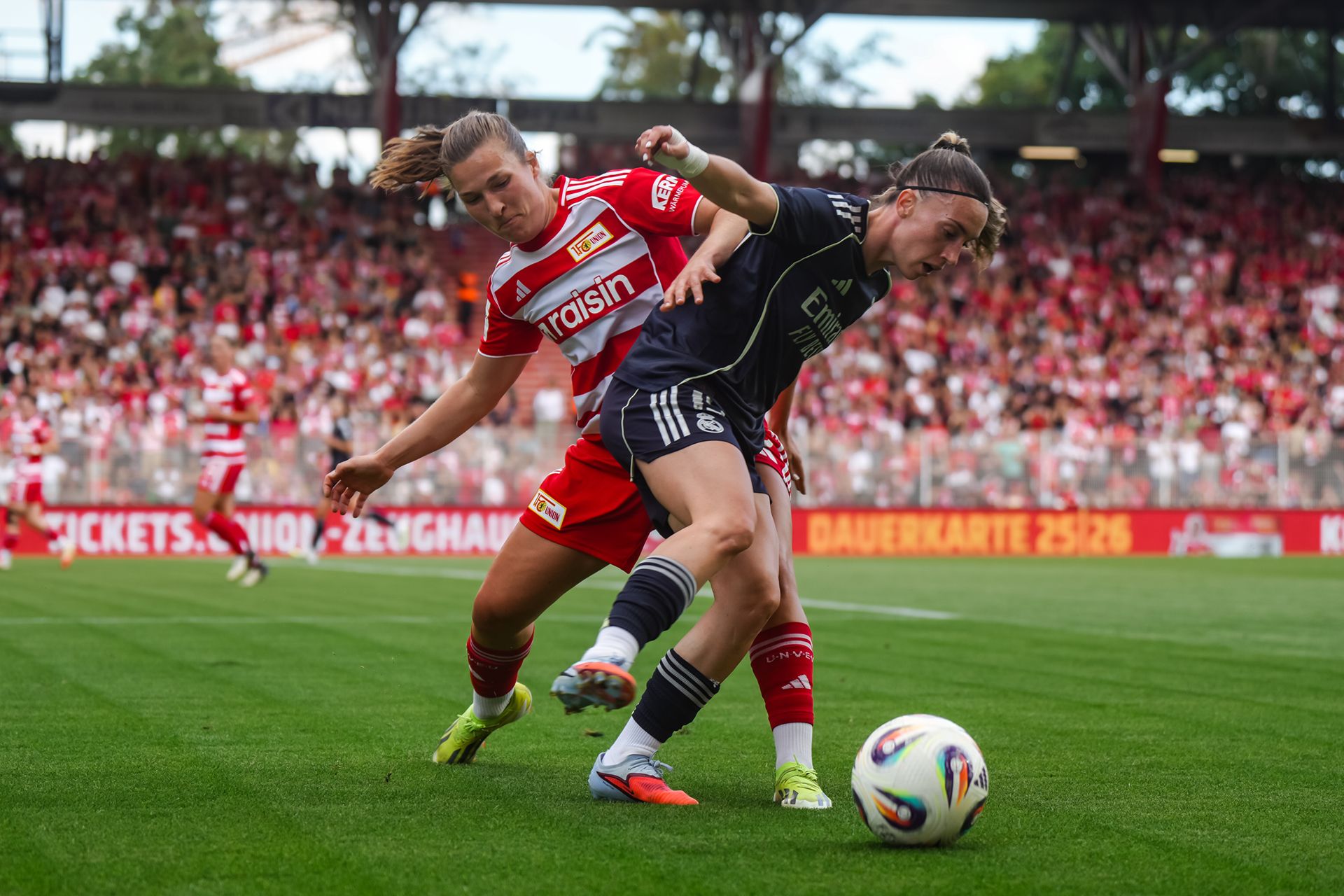 Zwei Fußballspielerinnen kämpfen um den Ball auf einem Spielfeld, während eine große Menschenmenge im Hintergrund zuschaut.