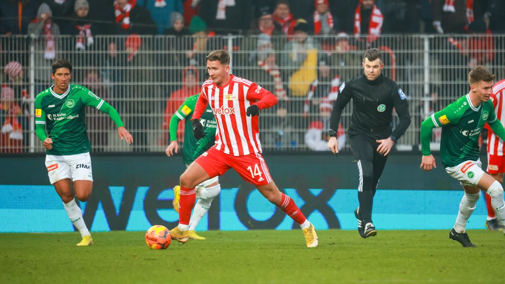 A soccer player in a red and white uniform dribbles the ball while two players in green try to stop him.