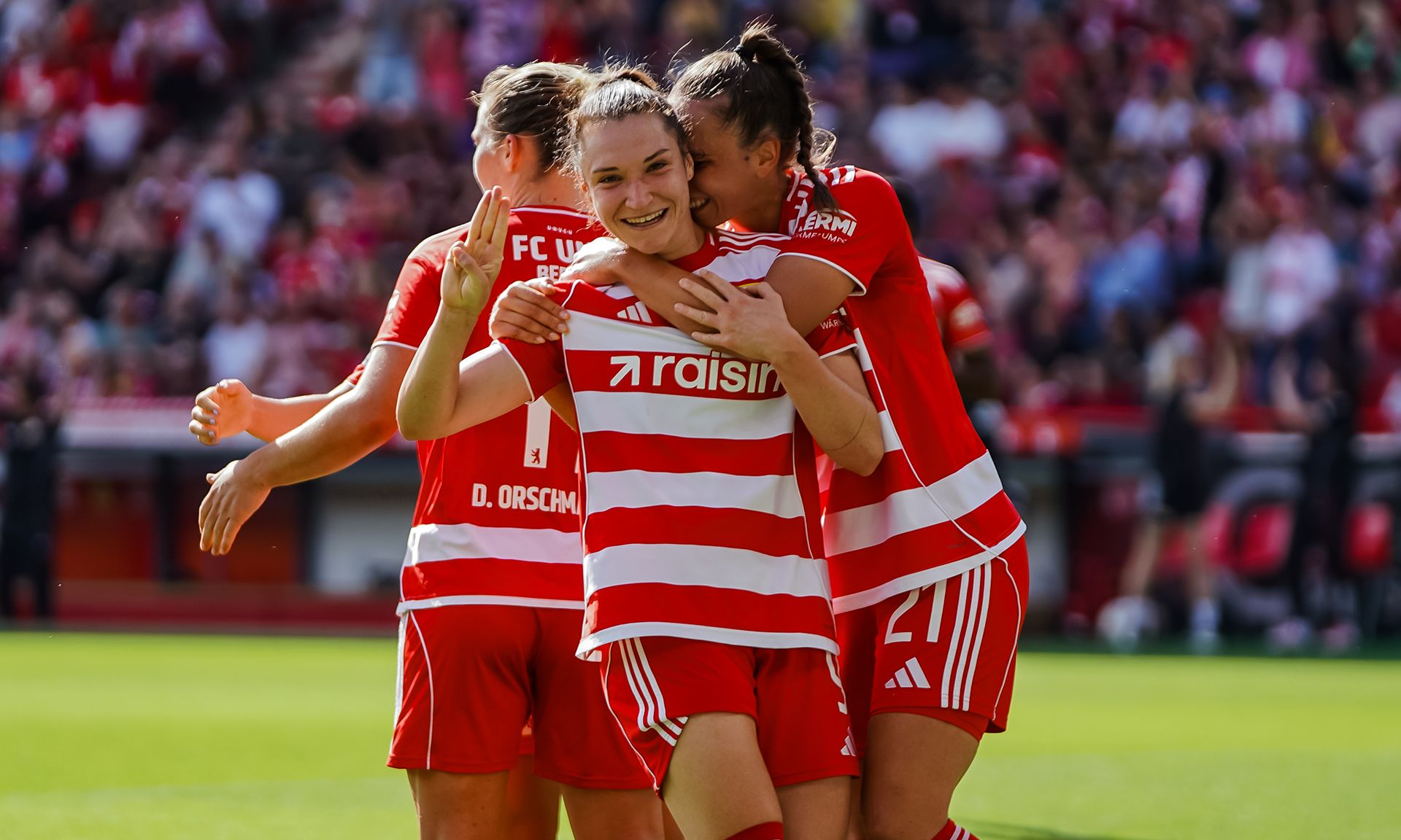 Eine Fußballmannschaft feiert einen Torerfolg auf dem Spielfeld, Spielerinnen umarmen sich vor jubelndem Publikum.