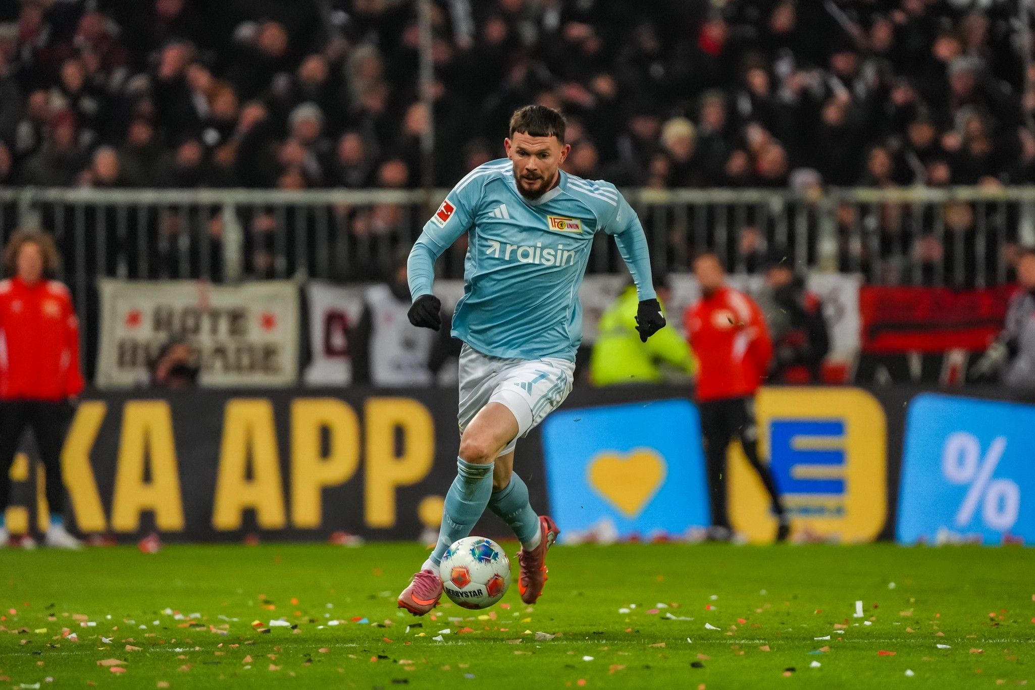 Spieler in hellblauem Trikot sprintet über das Spielfeld, umgeben von jubelnden Fans und buntem Konfetti.