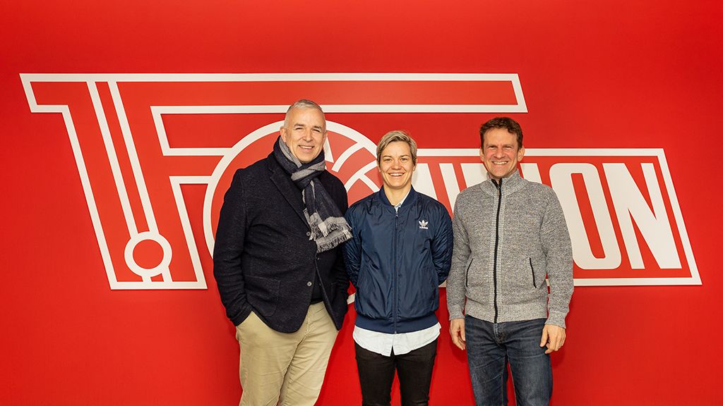 Three people are standing in front of a red wall with the FC Union Berlin logo.