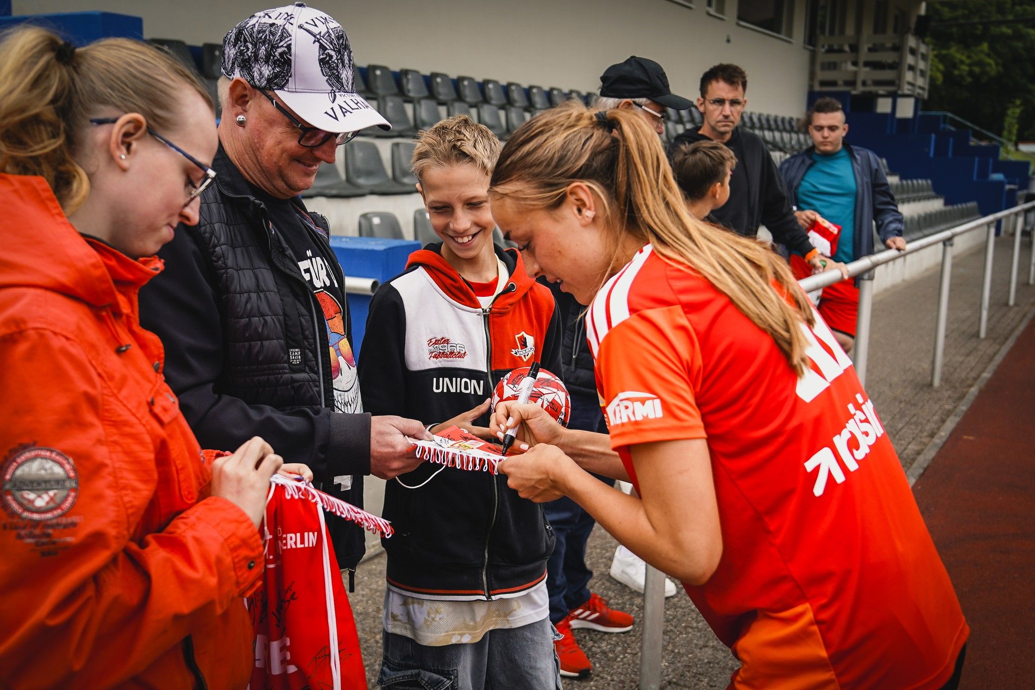 Eine Fußballspielerin signiert Trikots für Fans, darunter Kinder und Erwachsene, an einem Sportplatz.