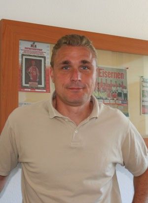 Ein Mann mit blonden Haaren steht lächelnd vor einem Holzrahmen mit Bildern und Zeitungsartikeln im Hintergrund.