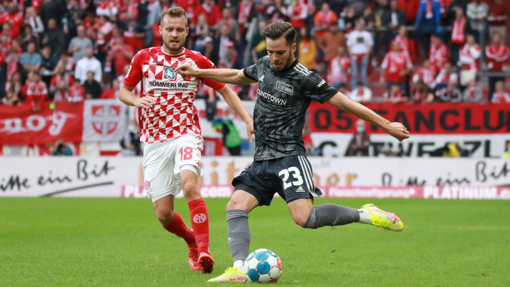 Two football players on the field, one kicking the ball while the other chases it. In the background, a cheering crowd.