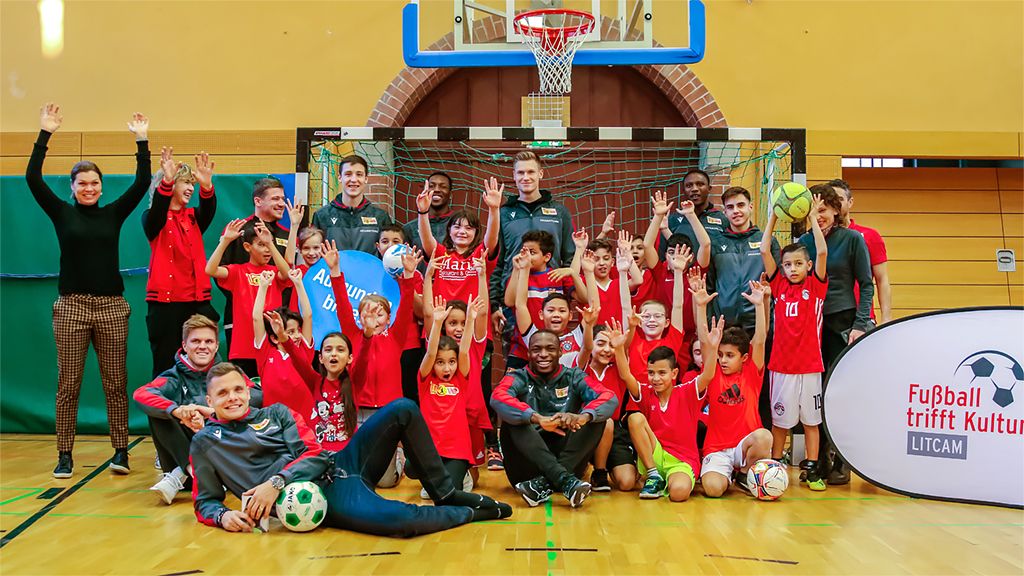 Gruppenshot von Kindern in roten Trikots mit Trainern in sportlicher Kleidung, die in einer Turnhalle posieren und die Hände heben.