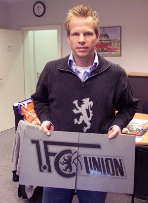 Mann hält ein Schild mit dem Logo des 1. FC Union Berlin in einem Büroraum. Im Hintergrund ein Tisch und ein Bild an der Wand.