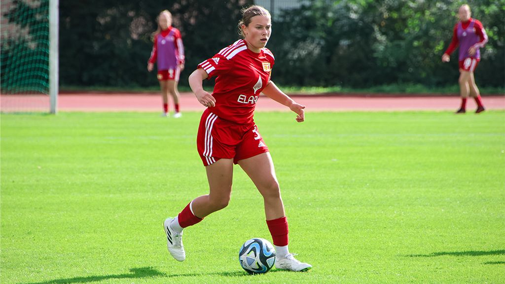 Eine Fußballspielerin in rotem Trikot dribbelt den Ball auf einem grünen Rasenplatz. Im Hintergrund sind weitere Spielerinnen sichtbar.