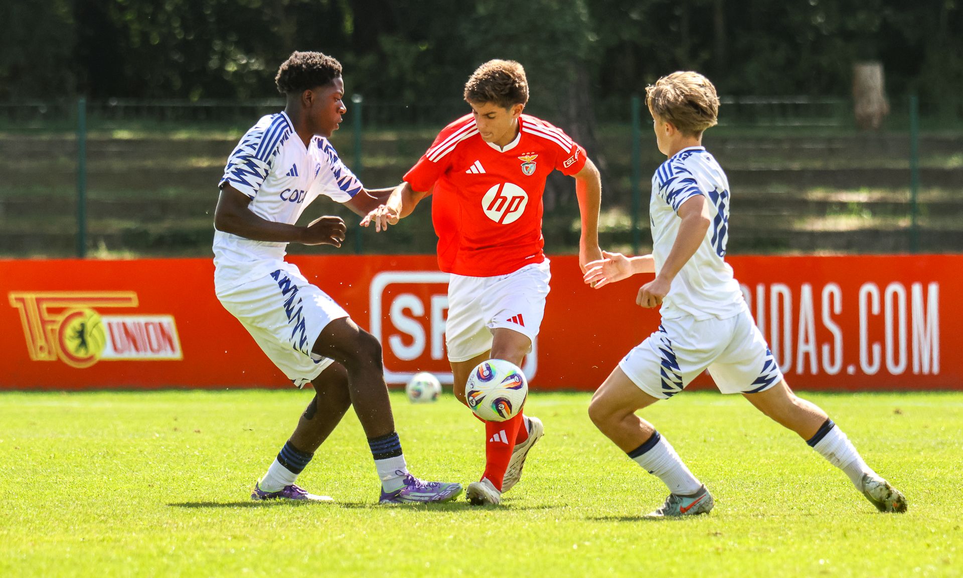 Ein junger Fußballspieler in rotem Trikot umdribbelt zwei Gegenspieler in weißen Trikots auf einem grünen Rasenfeld.