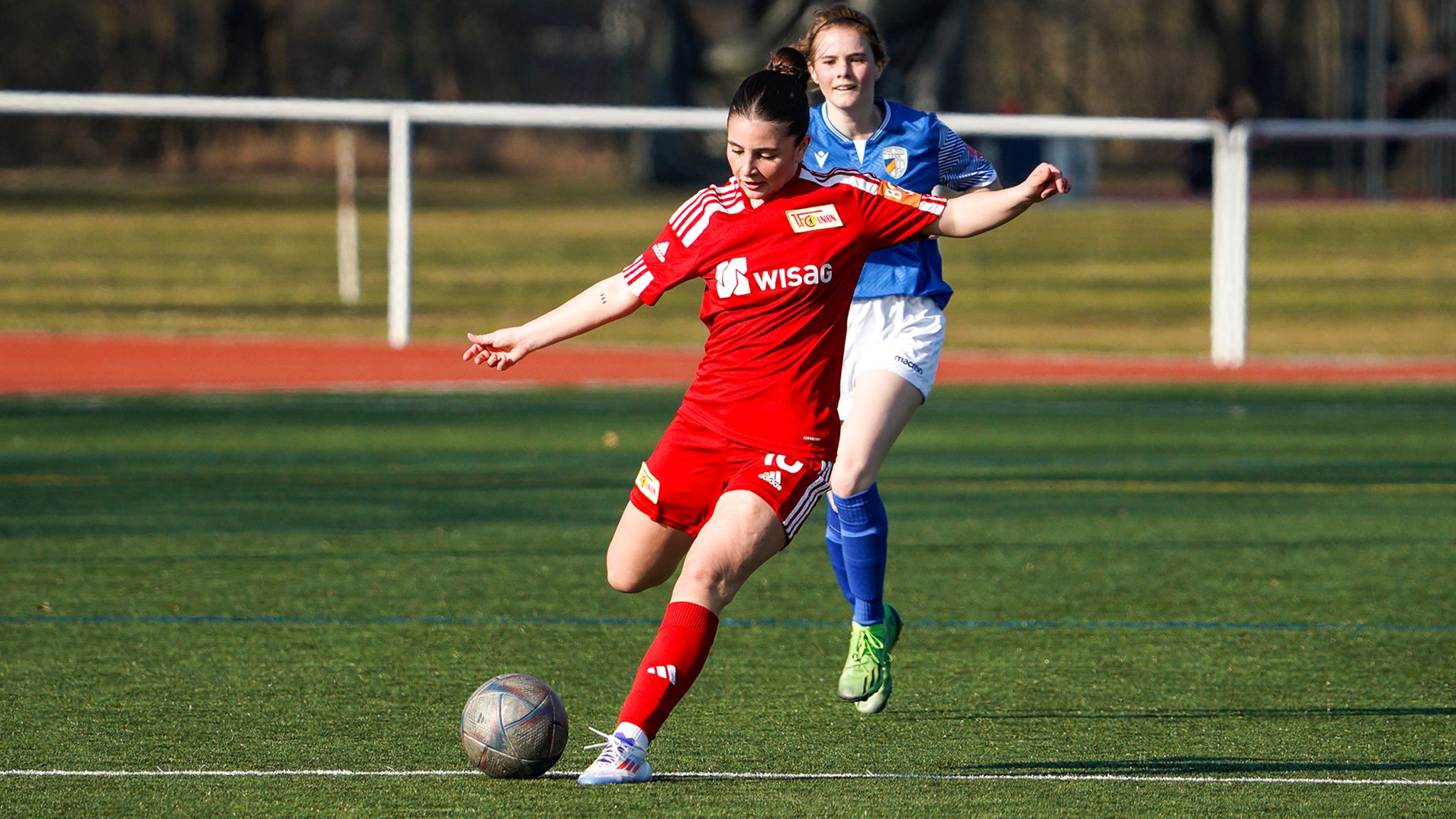 Eine Spielerin in rotem Fußballtrikot schießt einen Ball, während eine Gegnerin in blauem Trikot näher kommt.