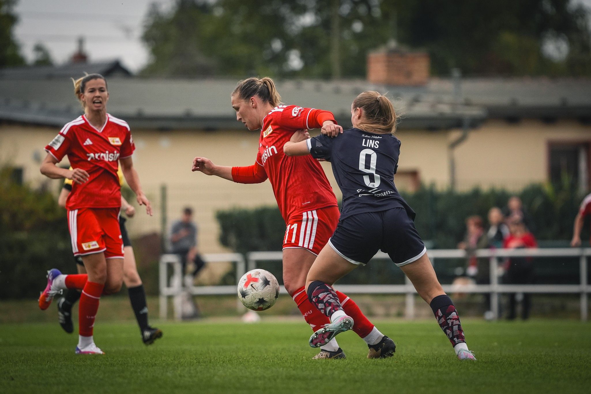 Spielszene im Frauenfußball mit zwei Spielerinnen im Zweikampf. Eine Spielerin in rotem Trikot wird von einer anderen in schwarzem Trikot behindert.