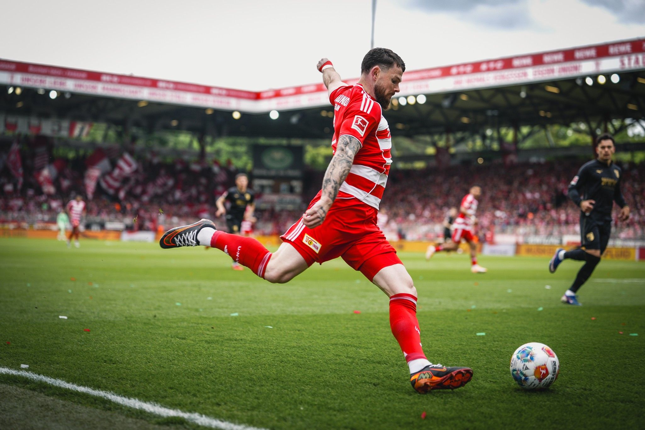 Ein Spieler in roter Fußballausrüstung führt einen Schuss auf dem Spielfeld aus, im Hintergrund Fans und eine Stadionatmosphäre.