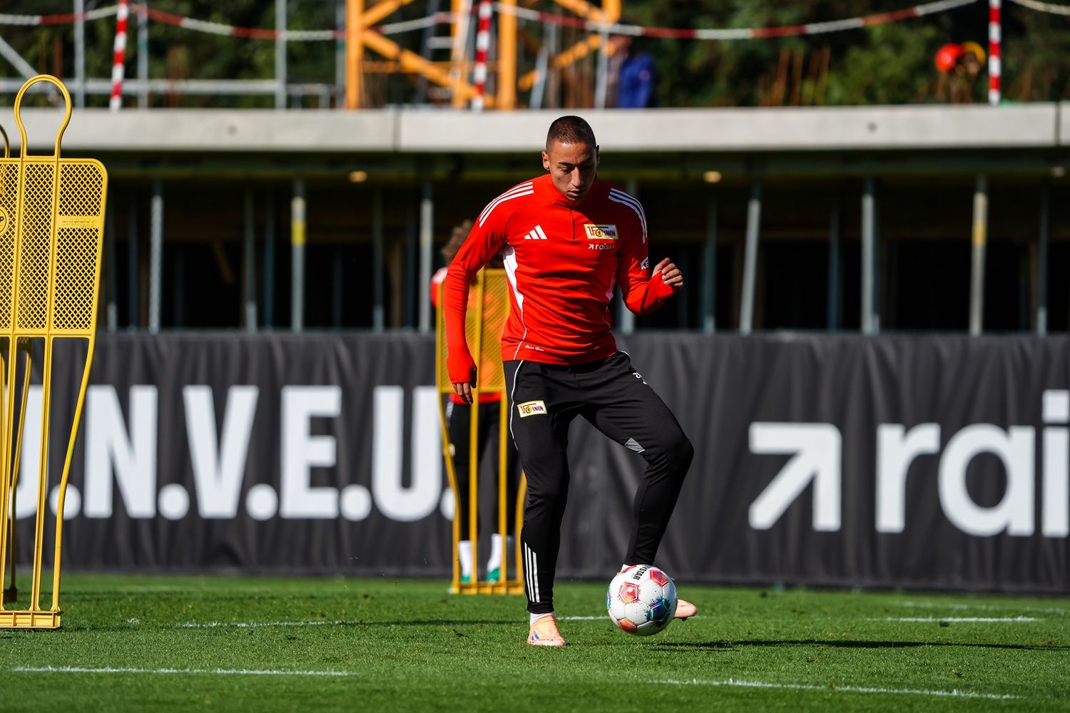 Ein Fußballspieler in rotem Trikot trainiert auf einem Platz mit Markierungshilfen und einer Trainingseinheit im Hintergrund.