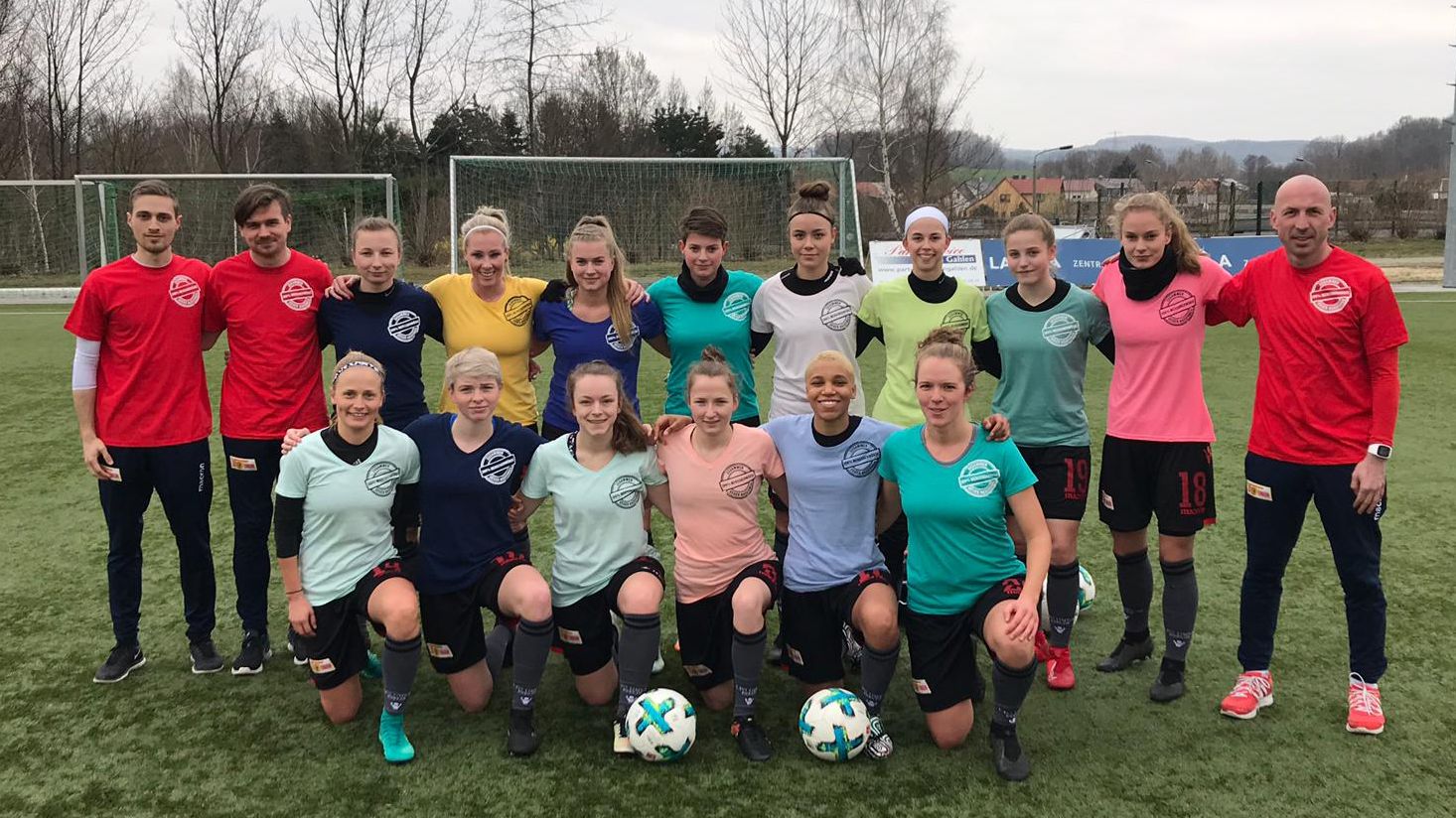 Eine Gruppe von 18 Fußballspielerinnen und -trainern in bunten Trikots steht auf einem Trainingsfeld.