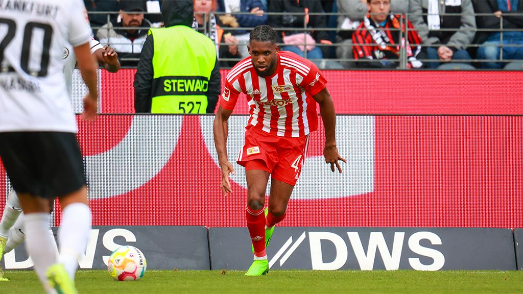 A football player in a red jersey runs with the ball on the field while spectators watch in the background.