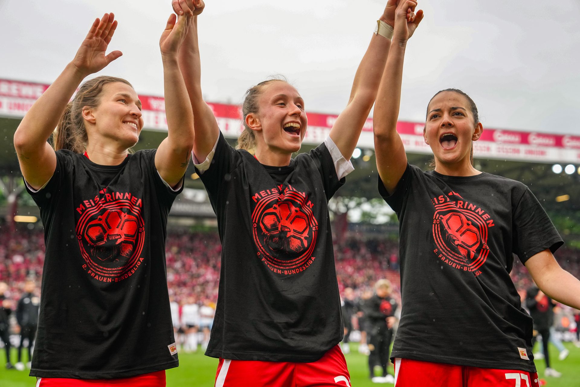 Drei Fußballspielerinnen feiern mit erhobenen Händen, tragen schwarze T-Shirts mit einem roten Fußballmotiv und grinsen in die Kamera.