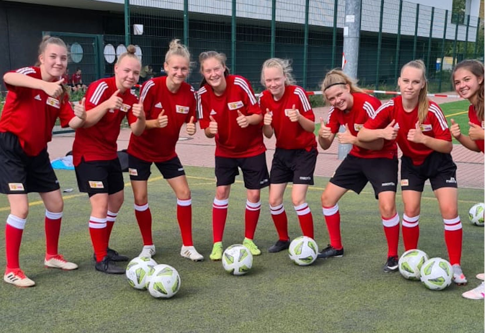 Eine Gruppe von neun lächelnden Fußballspielerinnen in roten Trikots gibt mit Daumen hoch ein positives Zeichen auf dem Spielfeld.