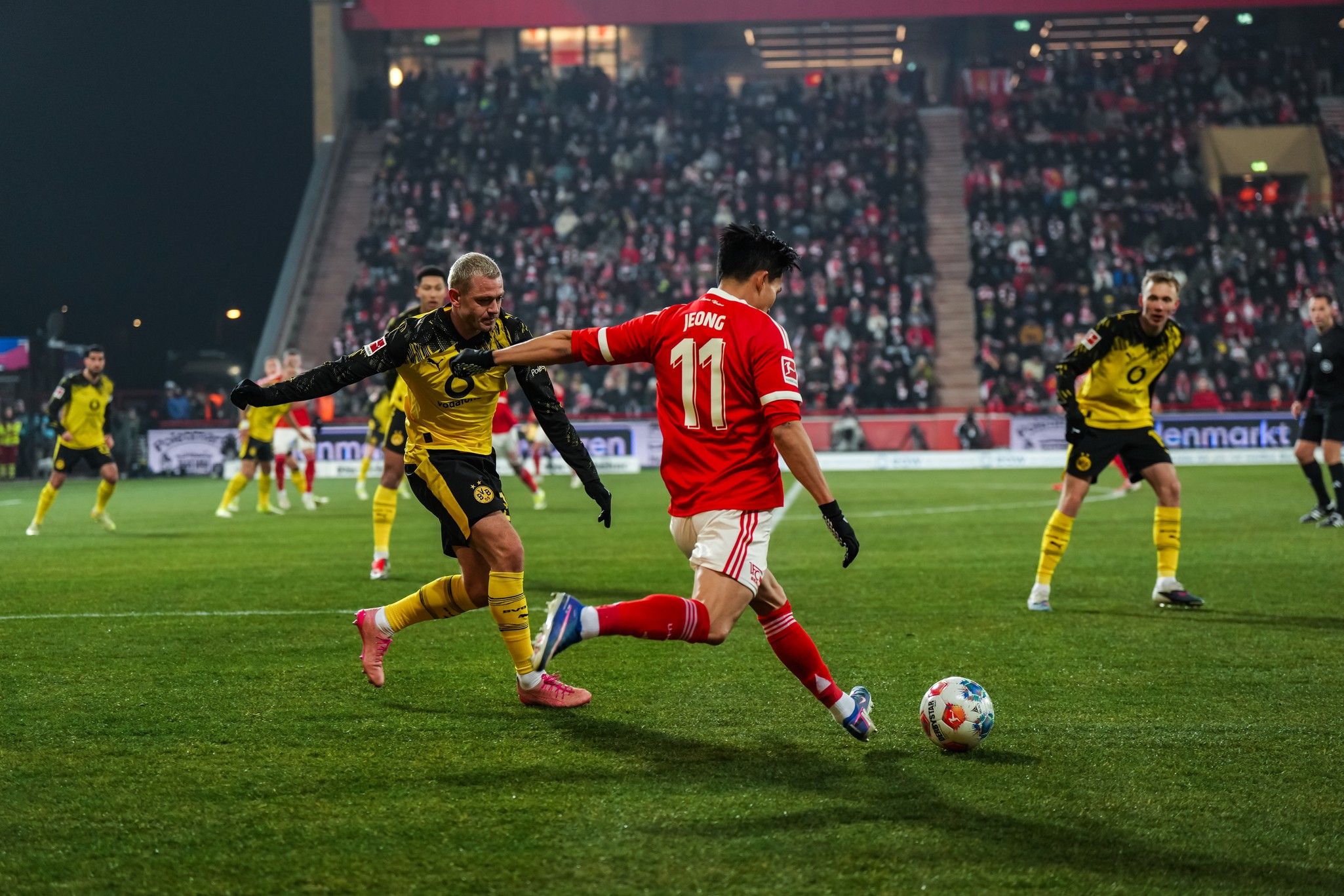 Spielszene mit einem Fußballspieler in rotem Trikot, der den Ball dribbelt, während ein gegnerischer Spieler in gelbem Trikot ihn verfolgt.