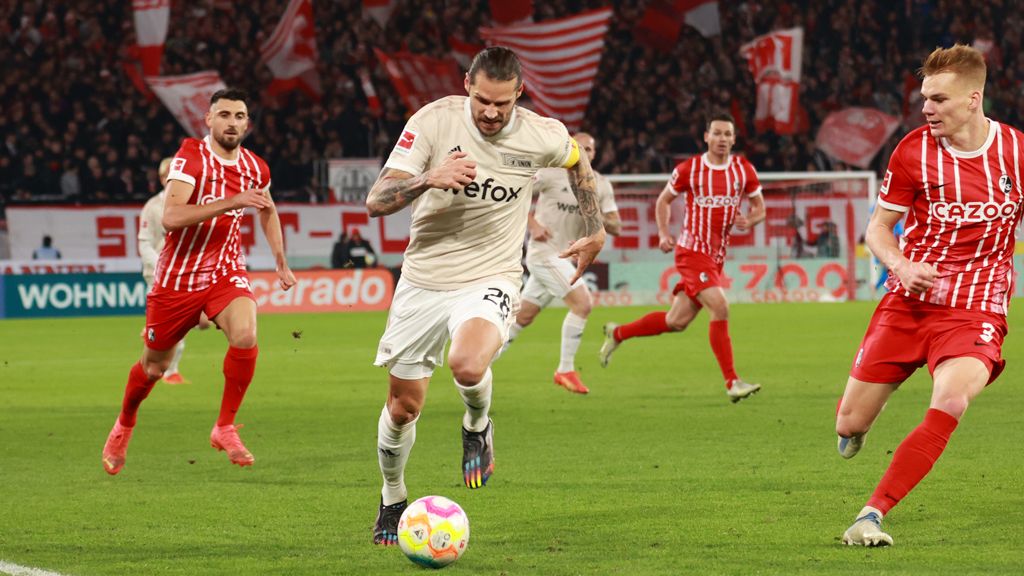 A soccer player in a light uniform is dribbling the ball while players in red jerseys seem to be chasing him.