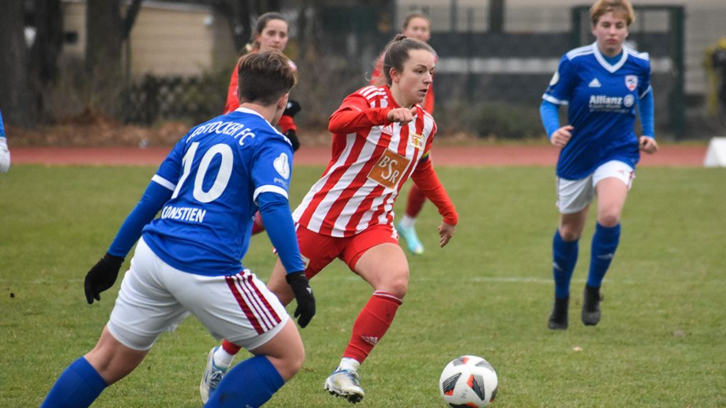 Eine Spielerin in rot-weiß gestreifter Uniform dribbelt den Ball, während zwei Gegnerinnen in blauen Trikots versuchen, ihn zu erreichen.