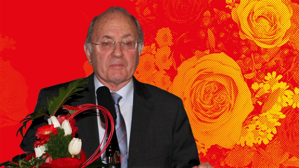 Ein Mann in Anzug steht vor einem Mikrofon, umgeben von Blumenarrangements, vor einem farbenfrohen, floralem Hintergrund.