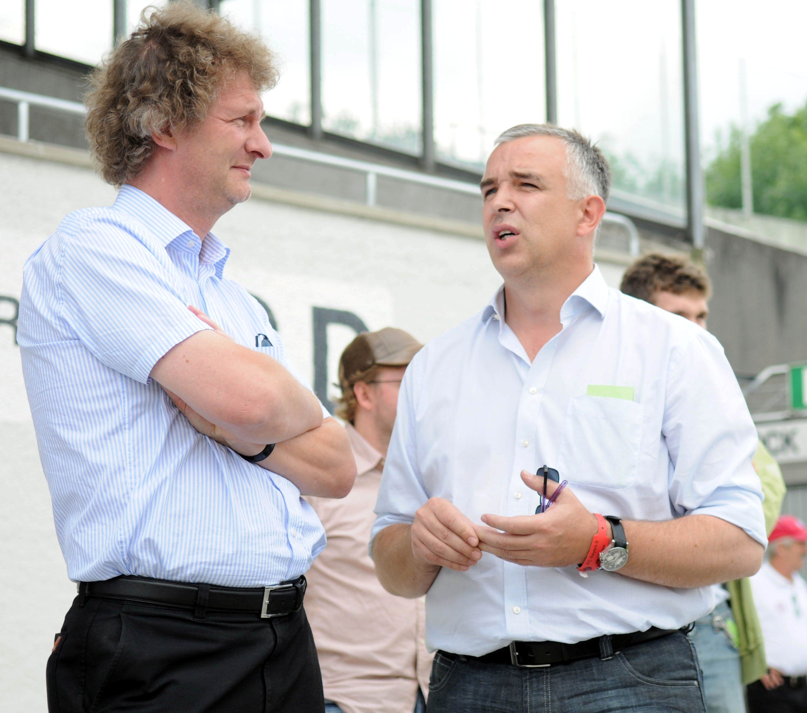 Zwei Männer im Gespräch, einer mit lockigem Haar und Hemd, der andere mit kurzem Haar und Brille, im Freien vor einem Sportgelände.
