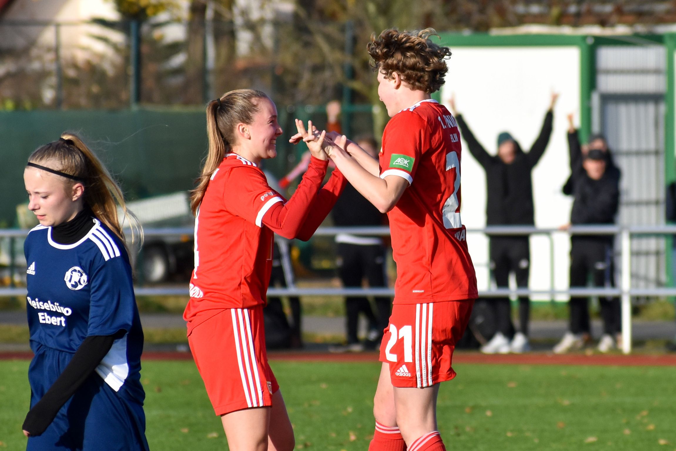 Zwei jubelnde Fußballspielerinnen in roten Trikots umarmen sich auf dem Spielfeld, während eine weitere Spielerin in Blau zusieht.