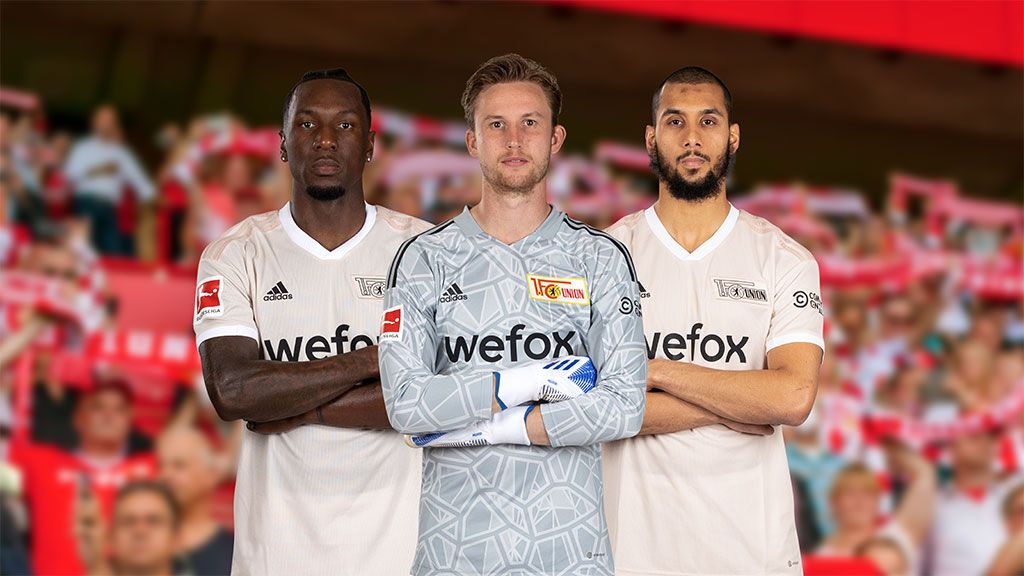 Drei Fußballspieler in weißen Trikots stehen zusammen, mit Fans im Hintergrund, die Schals halten.